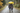 A couple walks away from the viewer on a wet gravel path during an autumn rain shower, sharing a bright yellow umbrella. The man, in a blue and red plaid shirt and jeans, has his arm around the woman, who wears a purple and black plaid jacket and dark pants; both are wearing boots. The scene is set amidst vibrant green and yellow autumn trees, with visible raindrops falling, creating a cozy and intimate atmosphere.