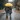 A couple walks away from the viewer down a wet asphalt road on a rainy day, holding each other close. The taller person on the left wears a dark plaid shirt and blue jeans, with an arm around the shorter person on the right, who wears a red plaid coat and blue jeans. They are both sheltered by a brightly colored yellow umbrella, with visible raindrops falling and trees with green and autumnal foliage lining the roadside.