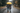A couple walks away from the viewer down a wet asphalt road on a rainy day, holding each other close. The taller person on the left wears a dark plaid shirt and blue jeans, with an arm around the shorter person on the right, who wears a red plaid coat and blue jeans. They are both sheltered by a brightly colored yellow umbrella, with visible raindrops falling and trees with green and autumnal foliage lining the roadside.