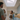 A candid medium shot of a family of four in a bright, modern kitchen. A smiling woman in a blue denim shirt holds papers and looks towards a bearded man in a green t-shirt, who has his back to the viewer and holds a young child. Another small child, also with their back to the viewer, stands between the woman and man. The kitchen features light blue cabinets, wooden countertops, a white subway tile backsplash, and a prominent skylight overhead, indicating a cheerful and well-lit home environment.