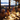 A diverse group of adults and seniors are seated around a beautifully set table at an outdoor dinner party during twilight. The scene is illuminated by warm string lights and candles, with guests laughing and enjoying their food and wine. Blurred city lights twinkle in the background, creating a festive and inviting atmosphere.