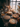A high-angle view shows a wooden table laden with several pies of various flavors, sitting in front of a sunlit window. The pies are arranged artfully with pumpkins, dried flowers, and lit candles. The scene evokes a feeling of warmth, abundance, and autumn.