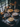 A dimly lit, eye-level still life photograph features five homemade pies arranged on a rustic wooden table. The pies have different fillings and crust designs. A window provides natural light, and fall decorations like pumpkins, foliage, and pinecones are scattered around the pies. A gray cloth is partially underneath the pies and along the table's edge.