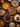 A top-down view of a collection of various pies arranged on a dark wooden surface. The pies include pumpkin, pecan, apple, and cranberry, along with other assortments. Scattered around the pies are autumn leaves, small pumpkins, pine cones, and bundles of cinnamon sticks, evoking a warm, autumnal, and festive holiday atmosphere.