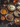 A high-angle shot captures a festive Thanksgiving dessert display. Several pies including pumpkin, pecan, apple, and a cream-topped pie sit on metal plates atop a dark, textured surface. Decorative gourds, pinecones, nuts, and fall foliage surround the pies, with a vintage-style teapot peeking from the background.