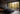 A luxurious modern bedroom features a large, dark gray upholstered bed with multiple pillows and a blanket, positioned against a dark brown vertical wood-paneled wall. Floor-to-ceiling windows dominate the wall opposite the bed, offering a stunning view of an autumnal forest filled with vibrant yellow and orange trees. Outside on a small balcony, a dark armchair and a round table with mugs sit, providing a cozy spot to enjoy the natural scenery.