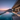 A wide-angle photograph taken at dusk of a luxurious modern villa with an infinity pool. The pool's deep blue water reflects the gradient sunset sky, which transitions from deep blue to purple and soft pink. The villa features a large concrete roof, an illuminated open-plan interior with a dining table, and an outdoor lounge sofa on a light-colored patio. Beyond the pool and glass railing, a vast ocean meets a sprawling city coastline, illuminated with numerous lights, all beneath a prominent dark mountain.