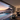 A wide shot captures a luxurious modern villa at dusk, showcasing an open-concept living room with a dining area, and an outdoor infinity pool. Beyond the pool, a panoramic view reveals a coastal city with twinkling lights, the vast ocean, and a prominent mountain peak under a vibrant pink and orange sunset sky. The interior is warmly lit, contrasting with the cool blues of the pool and the dusky outdoor hues, creating an inviting and serene atmosphere.