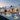 A breathtaking exterior view of a modern luxury villa at twilight, featuring a sleek, white facade with large glass windows and warm interior lights. A tranquil rectangular swimming pool with integrated lighting sits in front, surrounded by a manicured lawn, lush landscaping, and a stylish patio with an outdoor sectional sofa. The scene is illuminated by ambient warm lights from the house and cool blue hues of the dusk sky.