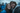 A close-up portrait of a young man and woman smiling happily and looking directly at the camera, sharing a clear umbrella covered in raindrops. The woman on the left wears a bright blue rain jacket and has long wet brown hair. The man on the right has a beard, wet brown hair, and wears a grey hoodie under a dark rain jacket. Blurred figures in similar blue jackets are visible in the background, suggesting an outdoor event on a rainy day.