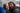 A close-up portrait of a happy young couple embracing under a clear umbrella during a rainy day. The woman on the left wears a bright blue rain jacket and smiles broadly, while the man on the right, with long dark hair and a beard, also smiles, with rain droplets visible on their hair and faces. The background is softly blurred, showing hints of other people and umbrellas.