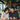 A happy, middle-aged East Asian couple is seated at a wooden table in a cafe, both smiling broadly. The man on the right, wearing a dark gray sweater, holds a black smartphone, which both are looking at intently, while the woman on the left, in a beige knitted sweater, holds a light gray mug. The background features blurred green potted plants on wooden shelves and bright windows, indicating a cozy, natural light-filled setting.
