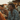 A young Black man with dreadlocks smiles brightly, looking left, while seated at an outdoor wooden table with friends during golden hour. He is wearing a blue paisley-patterned long-sleeve shirt and brown corduroy pants. Slices of pizza are visible on the table, and other blurry figures and buildings are in the soft, warm background, suggesting a casual social gathering.
