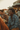 A young Black man with dreadlocks smiles brightly, looking left, while seated at an outdoor wooden table with friends during golden hour. He is wearing a blue paisley-patterned long-sleeve shirt and brown corduroy pants. Slices of pizza are visible on the table, and other blurry figures and buildings are in the soft, warm background, suggesting a casual social gathering.