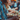 A close-up vertical shot shows a smiling Black man in profile, facing right, holding a paper tray with a colorful taco. He wears a blue patterned shirt with an intricate design, and his joyful expression reveals white teeth. In the soft-focus background, other people are visible, also eating, creating a vibrant, social outdoor scene with warm bokeh lights.