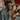 A young Black man with curly dark hair and a gold chain laughs heartily, holding a taco in his left hand, filled with fresh vegetables. He wears a blue and teal paisley-patterned shirt. Behind him, two other young people are smiling and blurred, also holding tacos, against a vibrantly colored background with red and teal architectural elements, conveying a lively and joyful atmosphere.