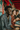A young Black man with curly dark hair and a gold chain laughs heartily, holding a taco in his left hand, filled with fresh vegetables. He wears a blue and teal paisley-patterned shirt. Behind him, two other young people are smiling and blurred, also holding tacos, against a vibrantly colored background with red and teal architectural elements, conveying a lively and joyful atmosphere.