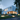 A photorealistic image of a minimalist, two-story modern white house at dusk, nestled within a dense forest of green trees and lush grass. Warm yellow light shines from the large windows, illuminating a serene interior, while ground lights highlight a path of stepping stones and large mossy rocks in the foreground. Distant mountains are visible under a soft blue sky, completing the tranquil scene.