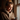 A young woman with light brown hair tied in a messy bun is illuminated by warm natural light from a window on her right. She wears a beige, textured sherpa hoodie and smiles softly while looking down, holding a dark smartphone in her left hand and a white coffee cup in her right. The background is softly blurred and darker indoors.
