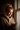 A young woman with light brown hair tied in a messy bun is illuminated by warm natural light from a window on her right. She wears a beige, textured sherpa hoodie and smiles softly while looking down, holding a dark smartphone in her left hand and a white coffee cup in her right. The background is softly blurred and darker indoors.