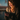 A young woman with curly, light brown hair smiles brightly while looking down at a smartphone held in her hands. She is illuminated by warm, golden sunlight streaming in from a window on the left, casting a beautiful glow on her face and hair, and wearing a textured light brown sweater. In her left hand, she also holds a mug, and the background through the window is a blurred blue and dark tone, indicating an indoor setting.