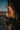 A young woman with curly, light brown hair smiles brightly while looking down at a smartphone held in her hands. She is illuminated by warm, golden sunlight streaming in from a window on the left, casting a beautiful glow on her face and hair, and wearing a textured light brown sweater. In her left hand, she also holds a mug, and the background through the window is a blurred blue and dark tone, indicating an indoor setting.