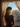 A full-length vertical shot from behind shows a person, likely a woman, with long, wavy blonde hair standing by a window. She is silhouetted against a bright, sun-drenched sky, with warm golden light creating a beautiful rim light on her hair and shoulders, and a sun flare obscuring part of her face. Beyond the sheer, patterned curtains framing the window, a clear blue sky, distant mountains, and an urban landscape of houses are visible.