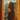 A young woman stands in profile, silhouetted against a bright window, looking out over a city and distant mountains under a blue sky. Golden sunlight streams in from behind her, beautifully illuminating her long, wavy blonde hair and the orange curtains framing the window. She is wearing light-colored loungewear, and her face is partially shadowed, conveying a pensive and calm mood.