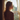 A young woman with long, wavy, light brown hair is seen in profile, standing by a window and looking out. Her shoulders and the back of her head are illuminated by strong golden sunlight coming through the window, creating a warm glow around her hair, while her body is largely in shadow. She wears a light-colored bralette, and the window is framed by sheer, light-colored curtains, with a blurred view of trees and a building visible outside.