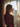 A young woman with long, wavy, light brown hair is seen in profile, standing by a window and looking out. Her shoulders and the back of her head are illuminated by strong golden sunlight coming through the window, creating a warm glow around her hair, while her body is largely in shadow. She wears a light-colored bralette, and the window is framed by sheer, light-colored curtains, with a blurred view of trees and a building visible outside.