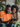 A happy Black man in an orange t-shirt and a Black woman in a beige knit sweater are sitting closely together, both smiling genuinely as they look at a dark gray digital tablet held by the man. They appear to be sharing a moment of engagement, possibly viewing content or browsing online, in a comfortable home environment with green houseplants and a blurred bookshelf in the background.