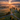 An aerial view of a vibrant rural landscape at sunrise. Golden light streams through a partly cloudy sky, illuminating a valley filled with lush green fields and soft morning fog. A paved country road intersects in the foreground, passing a white farmhouse and scattered trees, with distant rolling mountains visible on the horizon.