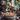 A baked apple pie with lattice crust sits on a stacked set of plates on a table with apples scattered around. In the background is a vase with light colored dahlias and orange leaves. There is a large window in the background showing trees outside and other objects scattered on the table.