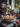 A baked apple pie with lattice crust sits on a stacked set of plates on a table with apples scattered around. In the background is a vase with light colored dahlias and orange leaves. There is a large window in the background showing trees outside and other objects scattered on the table.