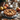 A beautifully arranged still life shows an apple pie as the focal point, set on a silver plate with a lace doily, topped with star anise and cinnamon. Three ripe apples sit nearby, along with cinnamon sticks and scattered dry leaves creating an autumnal ambiance. In the blurred background, a basket filled with fall foliage and a white ceramic teapot add depth to the composition.