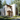 A visually striking, modern two-story house with off-white textured walls and warm wooden window frames is nestled deep within a dense, sunlit forest. The entrance features a wooden door and an overhead pergola, while the surrounding grounds are covered in wild green grass and small colorful flowers, leading to a path of irregular stone slabs. The scene conveys a sense of peaceful seclusion and contemporary design harmonious with nature.