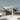 A high-quality architectural rendering of a luxurious, modern house with striking angular white roofs and extensive glass walls, set amidst a dense forest of tall trees under a clear, soft blue sky. Multi-tiered light concrete steps and stone-clad retaining walls lead up to the impressive entrance of the building, which blends contemporary design with its natural surroundings.