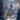 A stunning vertical shot of a snow-covered village at dusk or dawn. Charming stone houses with warm glowing windows and a tall, pointed stone tower are nestled among heavily frosted pine and bare trees. A glistening snowy path with subtle tracks leads through the scene, reflecting warm light from the distant buildings and decorated Christmas trees, all under a soft blue and purple sky with hints of pink.