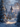 A stunning vertical shot of a snow-covered village at dusk or dawn. Charming stone houses with warm glowing windows and a tall, pointed stone tower are nestled among heavily frosted pine and bare trees. A glistening snowy path with subtle tracks leads through the scene, reflecting warm light from the distant buildings and decorated Christmas trees, all under a soft blue and purple sky with hints of pink.