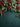 A top-down view of a Christmas border arrangement, featuring lush green evergreen branches, clusters of bright red berries, dark green holly leaves, natural brown pinecones, and small gold glittery baubles. These festive elements are artfully placed along the top and upper sides of the image, providing a decorative frame. The background is a dark teal, textured fabric, offering ample blank space in the lower section suitable for text overlays.