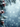 A vertical image showcasing snow-covered evergreen fir branches on the left side, featuring several rustic brown pine cones also dusted with white snow. The background is a soft, blurred expanse of icy blue with numerous white specks representing gently falling snow and soft bokeh lights. The overall scene evokes a peaceful and magical winter holiday atmosphere.