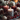 A close-up image showcasing three lit pillar candles as the centerpiece of a festive holiday arrangement. The central candle is gray with white speckles, flanked by a dark red speckled candle and a solid red candle, all emitting a warm glow. Surrounding the candles are frosted pinecones, bright red berries, small evergreen branches, and a decorative woven sphere, all lightly dusted with white powder on a dark gray textile, with blurred golden lights in the background.