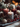 A close-up image showcasing three lit pillar candles as the centerpiece of a festive holiday arrangement. The central candle is gray with white speckles, flanked by a dark red speckled candle and a solid red candle, all emitting a warm glow. Surrounding the candles are frosted pinecones, bright red berries, small evergreen branches, and a decorative woven sphere, all lightly dusted with white powder on a dark gray textile, with blurred golden lights in the background.