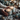 A close-up vertical shot shows a person's hands wearing a knitted sweater, gently holding two lit, textured gray pillar candles wrapped with twine. The candles and hands are surrounded by rustic winter decorations including frosted pinecones, evergreen branches, golden star anise, and scattered white glitter on a dark wooden surface with a soft gray fabric. A blurred background of warm, golden string lights creates a festive atmosphere.