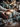 A close-up vertical shot shows a person's hands wearing a knitted sweater, gently holding two lit, textured gray pillar candles wrapped with twine. The candles and hands are surrounded by rustic winter decorations including frosted pinecones, evergreen branches, golden star anise, and scattered white glitter on a dark wooden surface with a soft gray fabric. A blurred background of warm, golden string lights creates a festive atmosphere.