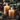 A vertical image featuring two lit, amber-colored pillar candles on a rustic gray-green plate. Around the base of the candles on the plate are frosted brown pinecones and sprigs of green evergreen. In the blurred background, more evergreen branches with scattered red berries and warm bokeh lights create a soft, festive ambiance against a dark wooden surface. The overall mood is warm, inviting, and perfect for the holiday season.