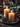 A vertical image featuring two lit, amber-colored pillar candles on a rustic gray-green plate. Around the base of the candles on the plate are frosted brown pinecones and sprigs of green evergreen. In the blurred background, more evergreen branches with scattered red berries and warm bokeh lights create a soft, festive ambiance against a dark wooden surface. The overall mood is warm, inviting, and perfect for the holiday season.