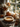A pumpkin pie decorated with whipped cream sits on a silver platter. Behind it, several pumpkins, pinecones, a candle in a jar, and a vase with flowers and leaves are arranged on a white windowsill in front of a window. The lighting is soft and natural, creating a warm, autumnal ambiance.
