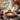 A pumpkin pie sits on a silver plate on a wooden surface, dusted with powdered sugar. Around it are white pumpkins, dried flowers in glass jars, pinecones, and a lace tablecloth. A window with blurred autumn foliage in the background provides soft, natural light.