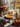 A brightly lit still life features a pumpkin pie on a cake stand in front of a window. The pie has a golden-orange filling and is decorated with small swirls of whipped cream around the edges. Arranged around the pie are a pumpkin, pine cones, candles, a vase of dried flowers, and a glass jar filled with dried nuts, creating a warm and inviting autumn scene.